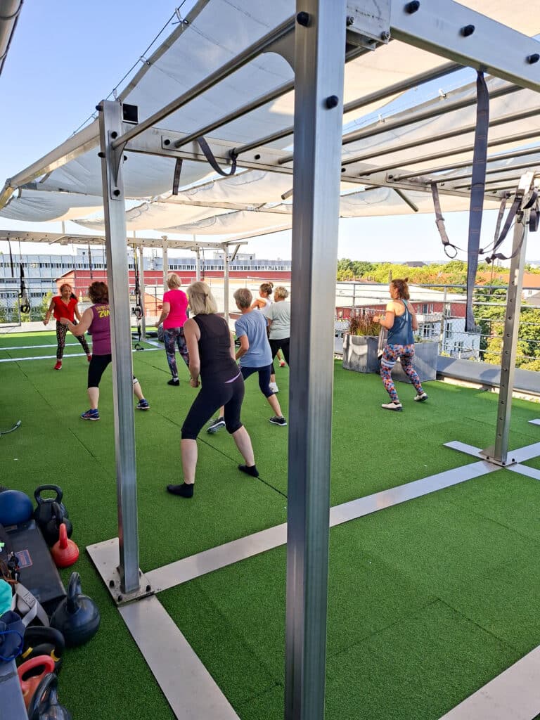 Kursgruppe trainiert auf der Dachterrasse von INJOYmed Dresden unter Sonnensegeln auf Kunstrasen.